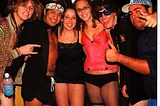 Six college students pose for a picture, all clad in scantily-dressed clothes. They are squished together and the room is bright, as if they are at a party.