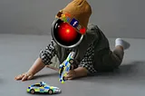 A toddler playing with toy cars. The cars are Irish police cars. The toddler’s head has been replaced with the menacing, glowing red eye of HAL9000 from Stanley Kubrick’s ‘2001: A Space Odyssey.’ The toddler’s knit cap is decorated with the logos for Apple, Google, Facebook and Tinder. Image: Cryteria (modified) https://commons.wikimedia.org/wiki/File:HAL9000.svg CC BY 3.0 https://creativecommons.org/licenses/by/3.0/deed.en