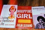 Three books of feminist theory laid on wooden background( Living A Feminist Life-Sarah Ahmed, Whipping Girl-Julia Serano, and Feminist Theory-bell hooks).