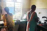 A kitchen with light streaming in from the far window. One housemaid is a the sink doing the dishes. Another is at the stove cooking lunch.