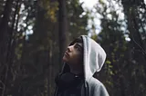 Teen in a gray hoodie gazing upward in a wooded area, looking thoughtful and introspective.