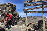 Trekker stands next to a rock face putting items in his backpack. Signboards tell climbers they are now at Gilman’s Point on Mount Kilimanjaro