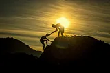 Twilight. Two hills. Two silhouettes on the hill in the foreground, one helping the other climbing to the top of the hill.