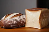 A close-up photograph shows two contrasting loaves of bread side by side on a smooth wooden surface. On the left, a round artisanal sourdough bread with a golden-brown crust displays an intricate pattern of scoring marks and a dusting of flour. On the right, a rectangular factory-produced white bread loaf wrapped in plastic is sliced into uniform pieces, showing soft, dense crumb structure. Both loaves are illuminated by soft directional lighting from above, emphasizing the textural differences