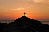 The image of a cross on a rocky shoreline at sunset.