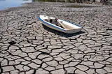 A small white boat stranded on a cracked and dry lakebed, surrounded by parched earth with deep fissures, symbolizing severe drought conditions and water scarcity