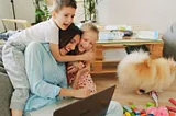 Mom on floor with laptop smiling while boy and girl hug her