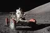 An astronaut drives a lunar rover across the rocky surface of the Moon, with hills in the background and a dark, starless sky above.