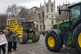 A road on which several tractors are going down with protestors holding up placards with messages on them.