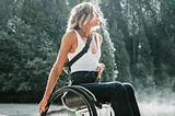 A model in a wheelchair poses in front of the forest. She’s wearing a white tank top and black pants.