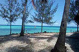 A red-colored sailboat just off the coast of Pemba — the sea is various shades of blue and there are trees growing from white sand in the foreground.