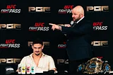 A man sits at a table with an announcer holding a microphone standing beside him and pointing.