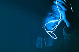 Person in a dark background with a cobalt blue light on their face, looking despondently at the camera.
