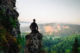 A person sitting on a rock ledge overlooking a lush forest and distant cliffs, symbolizing mindfulness and peaceful contemplation in a natural setting.