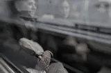 An old man’s wrinkled hands in sharp focus through the window of a passing train, the rest of the man and the person next to him blurred and streaked by the glass or motion