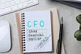 A notebook with the words ‘CFO Chief Financial Officer’ handwritten in bold colors, alongside a pen and keyboard, symbolizing financial leadership and strategy in the workplace. RAMS by Baz logo is featured, highlighting the brand’s focus on executive financial solutions.