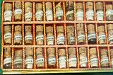 An antique wooden box containing multiple small glass vials with cork stoppers, each labeled with various substances in faded text, likely representing an old apothecary or medical collection.