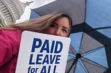 Woman with black and blue umbrella standing in front of US Capitol building holding a sign that reads “Paid Leave for All”