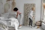 Two women using a cleaning schedule. One is making the bed while the other sweeps the floor
