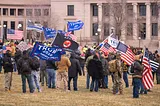 This is a group of protestors on a lawn outside a government building. Some have US flags, some have TRUMP 2020 flags, and a placard reads “STOP THE STEAL”