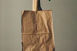 Paper grocery bag sitting on a stool, with a fist sticking up out of the bag, brandishing a clump of dark green cilantro