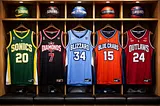 Fictitious jerseys of the Seattle Sonics, Las Vegas Diamonds, Buffalo Blizzard, Baltimore Blue Crabs, and Kansas City Outlaws hang from locker room lockers with matching team basketballs sitting above them.