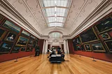 An art gallery room with high curved ceilings, parquet floor and old paintings in golden frames with a bench at the center to visitors to rest