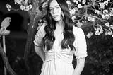 Girl in long white dress stands in front of a blossoming fruit tree in garden. Black and white photo.