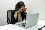 A photo of a woman seated at a table with a computer in front of her, holding her head in her hands.