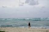 A woman looks out at a stormy ocean from the beach with shoes in her hand