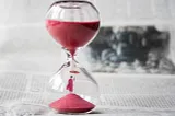 The picture shows an hour glass filled with red sand. A tiny man is at the bottom of the glass holding an umbrella over his head.