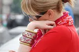 Blonde woman, hair tied back wears sunglasses, bangles and red clothes, looks thoughtful and writes in a journal