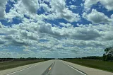 Color photo of open landscape, paved highway, and cloud-filled sky