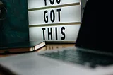 A picture of a small white sign with black letters on a desk next to a laptop. The sign says “YOU GOT THIS”.