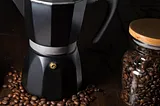 A black Moka pot coffee maker around coffee beans in a jar.