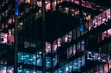 An image of windows of an office building at night with different color (purple, blue, pink) lights illuminating them from the inside.