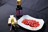 A neatly arranged plate of maguro (tuna) sashimi sits beside a bottle of nama shoyu (raw soy sauce) and a fresh wasabi root with a grater, all placed on a dark background.