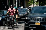 Three cyclists, one with a bike meant to carry a wheelchair-bound rider in the front, pass between a row of parked cars and moving cars. A large SUV is in the foreground.