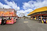 wide angle picture of a stall selling fair food, another stall on the right, and the ferris wheel in the background