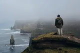 A man wearing a winter jacket standing on a grassy cliff. In the backgroud, the ocean is visible with steep cliffs rising up into the fog.