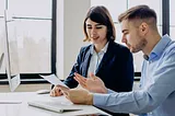 A photo of a young man and woman working together on a document at a computer workstation.