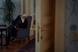 A view into a room through an open door of an older man resting his head on his hand, sitting in a chair beside a palm plant.