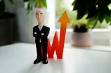 Photo of a doll of a smug-looking white man standing in front of an upward-trending line (as of stock price or financial projections)