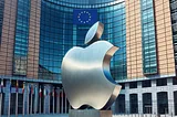 IMAGE: A Dall·E generated image of the European Union headquarters in Brussels, featuring a large, modern building with the EU flag prominently displayed, and in the front entrance patio, a metallic statue of the Apple logo