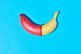 A banana lies on a bright blue background. The left half of the banana is covered with a red condom, creating a sharp contrast with the uncovered yellow right half. The image is brightly lit and has a playful, educational tone, likely symbolizing safe sex practices.