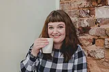 Author Lauren Aitchison standing in front of an exposed brick and white wall, wearing a checkered jumper, and holding a cup.