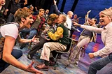 Two women frame a group of actors celebrating onstage as two characters kiss