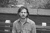 black-and-white photo of Adam Giannelli, a white man with a short beard and medium-length hair. He wears a plaid shirt and looks calmly at the camera.