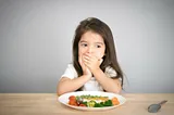 A young girl sitting at a dining table with food in front of her, covering her mouth or we call it the Mouth Shut Movement (MSM), with her hands despite the food being served