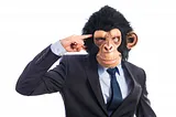 A man in a business suit and a full-head chimp mask pointing at his temple.